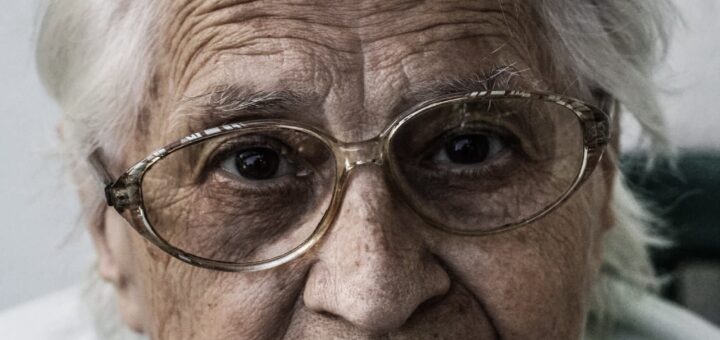 This vewry close image shows an older woman with white hair, a wrinkled forehead, and wire-rimmed glasses. She wears a white top with the shoulders barely visible. Her expression is stern with a tight mouth.