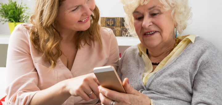 This indoor images shows a younger woman on the left. She has long, light brown hair and iw wearing a pink sweater. She's pointing to something on a smartphone that's held by an older woman with white hair. The older woman wears alight blue top with white trim. Both woman have slight smiles and are communicating about what they see with pleasure.