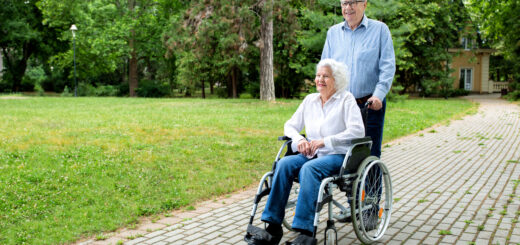 This outdoor image shows a park in the background. Int the foreground, a man is pushing a woman in a wheelchair along a brick path. The man has gray hair, glasses and is wearing a blue, long-sleeved shirt. The woman seated in the wheelchair has white hair, and is warhng a white, long-sleeved top and jeans.