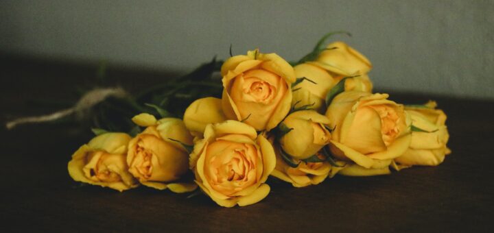 An indoor background shows a gray wall with a dark wood table in the foreground. Laying on the table is a bouquet of yellow roses seen from the top of their blooms.