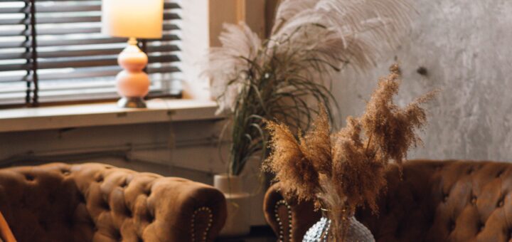 This indoor iage shows brown blinds in the background that are tilted open. One the window ledge sits a small lit lamp. There is a dramatic feather and dried plant arrangement in the corner by the window. A brown, tufted couch is visible on the left with a matching love seat on the right. A coffee table between olds a glass ornament.