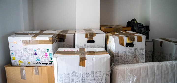 This indoor image shows two empty closets in the background. The walls are white. In the foreground we see piles of boxes taped with brown straping tape. Some boxes are white, others normal brown.