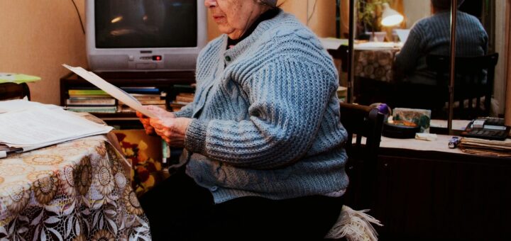 This indoor image shows an olde fashions room with a patterned oil-cloth table cloth. There is equipemnt hooked up around the woman sitting at the table with lots of cords evident. The woman is older with gray hair peaking out from a gray wool watch cap. She wears a bulky light-blue cardigan and is studying a paper in her hands.