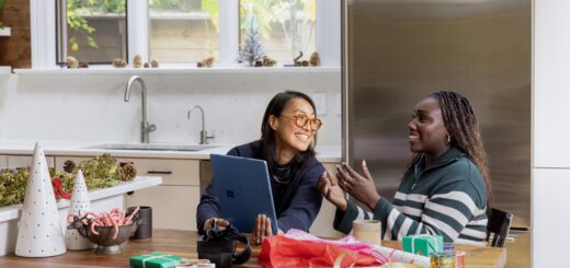 The scene for this image is a kitchen with large windows looking out onto trees. Two women sit next to each other at a table visiting and opening small gifts. One woman has light skin and long brown hair and she wears glasses. The other woma, on the right, has dark skin and dark hair pulled back. She wears a gray pullover sweater that lower down and over the arms has gray and white stripes.