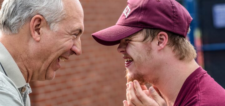 This outdoor image shows a dirt ball field in the background. In the foreground, we see the profile of an older man with gray hair and a receding hairline, with a broad smile as he pats a middle-school child on the shoulder. The child is wearing a maroon baseball cap and is smiling back in triumph as they receive congratulations.
