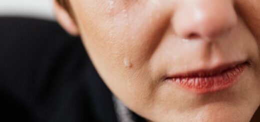 An extreme closeup of a the lower portion of a feminine face and black clad shoulder is all we see. We can see a portion of the ear and lower eye, half the nose, and half of the mouth that wears rose lipstick. Quiet tears stream down the cheek.
