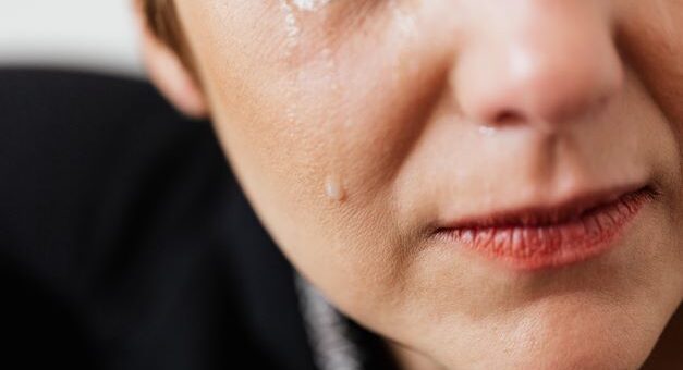 An extreme closeup of a the lower portion of a feminine face and black clad shoulder is all we see. We can see a portion of the ear and lower eye, half the nose, and half of the mouth that wears rose lipstick. Quiet tears stream down the cheek.