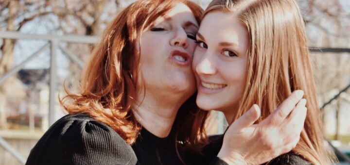 This outdoor image shows a spring tree flowering in the background against a blue sky. In the foreground, we see two women. Oe has long redish hair. Shes' wearing a black sweater and hs her head tilted back and to the said as she kisses a younger woman on the cheek. The younger woman is looking to the side but also up a little. She has longish light-brown hair and is smiling.