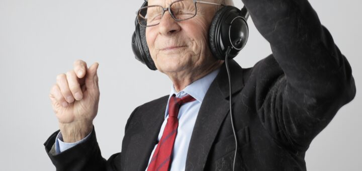 The background is a grayish wall. In the foreground is an older man with large headphones over his balk head. He's wearing a black suit, white chirt and red tie, and glasses on his face. His eyes are squeezed shut in concentration and he waves his arms as if directing an orchestra.
