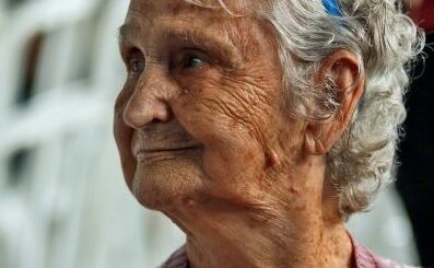 This image is outdoors with a blurred white background. IN the foreground, we see an older woman with short, curly gray hair wearing a dark blue headband. Her head is turned to profile and she wears a mauve and white top, probably a sweater but we only see the shoulders. Her expression is one of detrmination and appears negative.