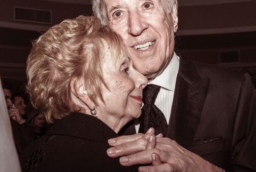 This sepia-toned image shows an older couple embracing. The woman has short brownish hair and a dark top. We only see her profile. The man is a bit taller. He has a receding hairline showing bray hair and is wearing a dark suit and tie with a whilte shirt. Both have medium toned skin and are wearing contented smiles.