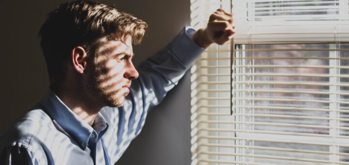 This image is inside a room with gray walls and with apinted trim. A middle-aged many is staring out the window through the open blinds with a thoughtful look on is face that is seen in profile. He has light brown, curly hair in a shorter cut, light skin, and wears a long-sleeved gray collared shirt. His arm is raised as he braces himself against the wall beside the window.