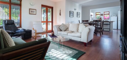 This indoor image shows a large white interior with a windowed exterior door. Several couches and other comfortable seating is visible as is a turquoise area rug. in the distance, we see a dining room and another room beyond that.