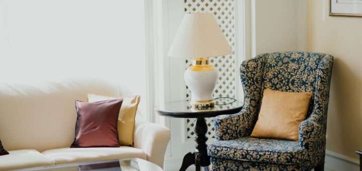 We view a nice interior of a sitting room. IN one corner is a blue print wing chair with a gold throw pillow. on the side is a round glass lamp table with a white reading lamp. Nex to that is a white couch. We see just on end that holds a red and a gold throw pillow. In front of that is a glass topped coffee table. There is whte lattice work wood behind.