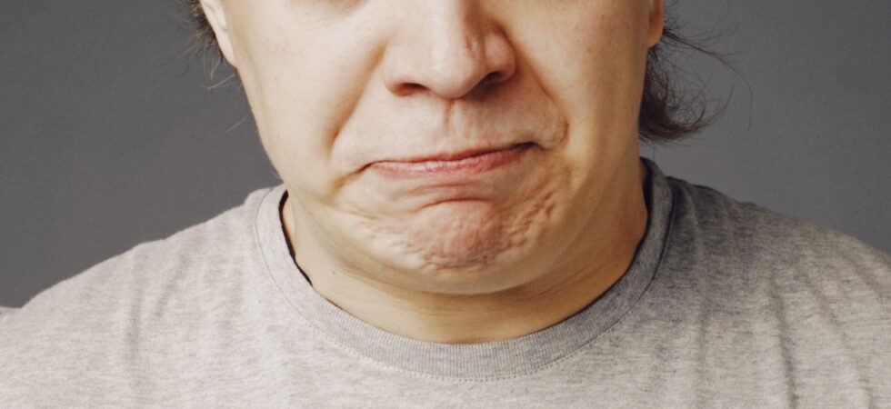 This indoor image shows a closeup of a man's face. He's middle aged with brown hair, whispy bangs, light skin and blue eyes which are widened with eyebrows raised in an exageraged look of surprise. Hes mouth shows bemusement or confusion. His clothing is plain so all we see is a beight tee shirt.