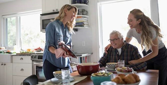 this indoor image shows a moder kitchen with white walls and cupboards in the background, though we see an open doorway. At a table, sits an older man with a receding hairline. His hair is white, and he wears dark rimmed glasses and a black and red buffalo plaid robe. A younger woman with long blonde hair, light skin, and causual blue tee with jeans is smiling as she hands the older man a plate. We see the arm of someone else around the shoulder of the older man.