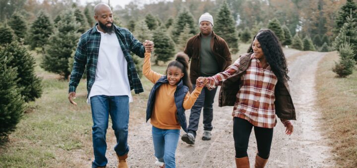 This image shows a multi-generational family walking joyfully down a country road. We see evergreens in the near background and other trees further back. The family in fore ground shows an adult male wiht dark skin wiearing an oversided white tee, a blue palid open flanner, jeans ad boots. He hold the uprased hand of a child who ears an orange lon-sleeved sweather dark blue scarf around shoulders, light blue jeans and boots. The child holds the other hand up to grabe the hand of a oman with long dark curly hair, a brown and white plaid, pullover sweater, jeans and boots. Behind them all is an older, white haired man wearing a brown jacket open over a blue shirt. He also wears jeans but with casual trainers.