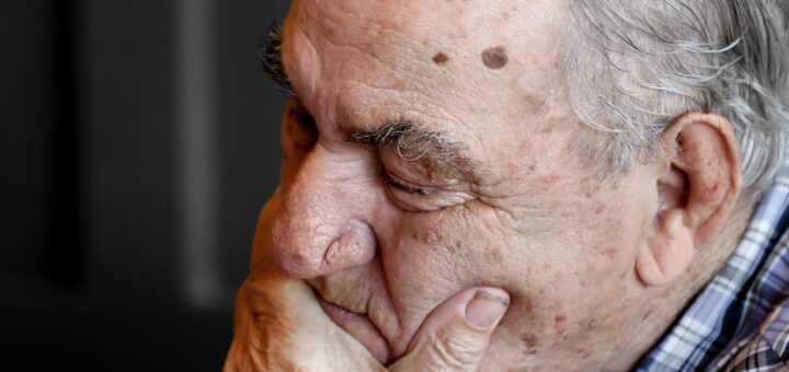 We see an image with a dark, textured background. In the foreground, we see an older man in profile. He has light skin, gray hair, and two moles on his temple. - one large and one small. He's holding his had over his mouth and is looking downward, either hin frustration or due to sleep.