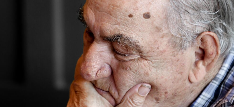 We see an image with a dark, textured background. In the foreground, we see an older man in profile. He has light skin, gray hair, and two moles on his temple. - one large and one small. He's holding his had over his mouth and is looking downward, either hin frustration or due to sleep.