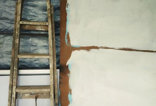 A tall painter's ladder rests agaist a wall that's been torn open to show the backing. The viewer sees unfinished patched plaster or drywall on the side.