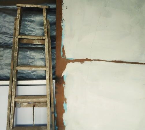 A tall painter's ladder rests agaist a wall that's been torn open to show the backing. The viewer sees unfinished patched plaster or drywall on the side.