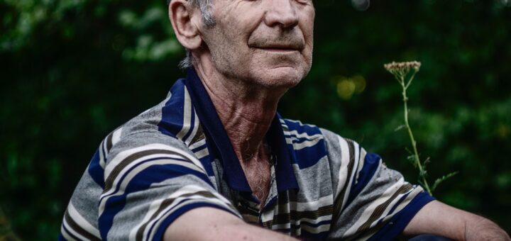 This outdoor scene shows thick bushes in the background. In the foreground, we see an older man who is bald in fromt but has gray hair along the sides. He sits on teh broud with his arms wrapped around his knees as he looks off into the distance. He's wearing a black, gray, and white striped golf-style shirt.