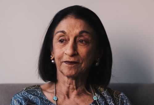 This indoor image shows a woman who looks to be of middle eastern heritage. She's got shoulder length black hair and is wearting a turquoise paisley top and turquoise enhanced necklace. Her expression is one of puzzlement.