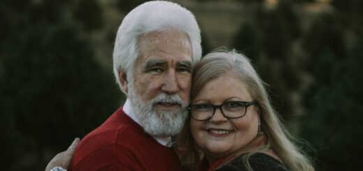 This image has a blurred outdoor background with pine trees. In the foreground we see an older man with very white hair and a trim beard and mustache. He wears a dark red jacket or sweater. Hugging in slightly from the side is a younger woman with very long, dark blonde hair and dark-rimmed rectangular glasses. She wears a very dark read garment that we see covering her arm that encircles the man.