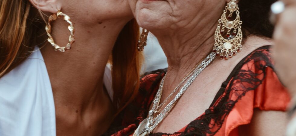This ismage is festive as if at a celebration. Two women are in the foreground. One is younger with long light-brown hair. She is seen in profile weathing a gold hoop earring and a white jacket as she offes a kiss to the cheek of an older woman. The older woman has died dark curly hair and also seen in semi-profile. She wears a large, ornate gold earring, and gold necklace and and orang and black ruffled top that oculd be worn off the shoulder. We see part of a bare arm.