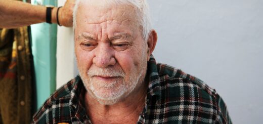We see a generic indoor background done in gray with green accent. An old man sits in a chair where we see a wood rail across the back by his head. The man has short, cropped white hair and a neatly trimmed white beard and mustache. He has medium toned skin and a mole on his cheek. He wears a dark plaid flannel and we see the rounded head of his cane leaning against his chest.