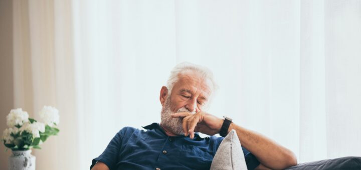 We see an older man in a chair with his head tiltled toward his hand. He seems to be sleeping. He has white hair and wears a dark blue short-sleeved shirt. There's a pillow propping him up on one side.