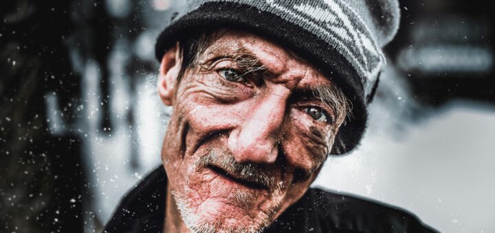 The outdoor background is gray and blurred. Int eh foreground we see an older man with a weather brown face and slightly crooked nose and white beard stubble. He's wearing a gray winter cap with a logo of some kind tipped up. HIs dark overcoat is barely visible.
