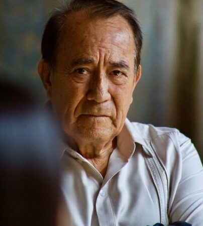 this indoor image has a blurred background. IN the foreground we see a man with clipped brown hair and a receeding hairline who wears a cream golf-style shirt with some muted stripes. He has brown skin. He wears an unhappy frown and he looks slightly toward the side.