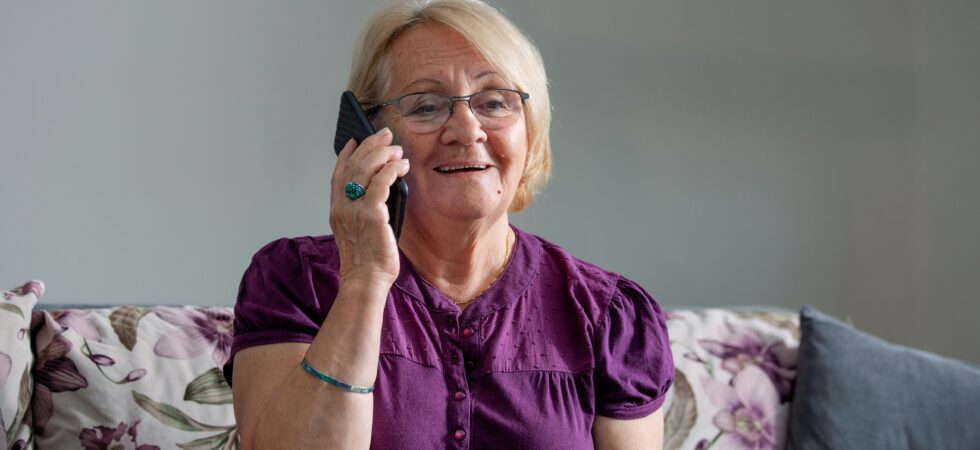 This indoor image shows a gray wall in the background. An older woman sits on a lavender and green print couch She has short, dyed blonde hair and wears a purple short-sleeved dress and glasses. She holds a smart phone to her ear and she listens to someone talking to her. Her expression is pleasant with a slight smile.