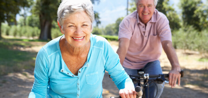 This outdoor image shows summer trees and sky in the background on a city street. In the foreground we see an older couple riding bikes. On the left is a woman with short gray hair wearing a light blue Henley top. Slghtly behind her on the right is a man with gray hair wearing a pink golf shirt. Both are smiling as they enjoy their outing.