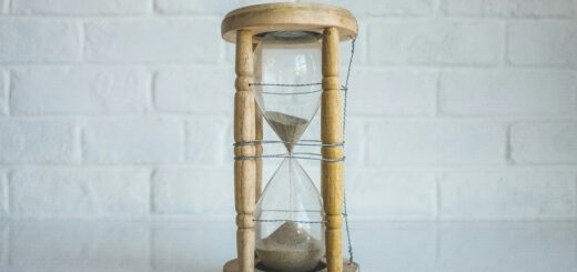 Image shows a white painted brick wall in the background. A classic hourglass sits on a white table. The hourglad is made from light wood and the sand is about 2/3 of the way through.