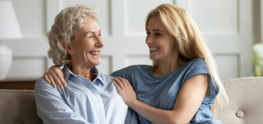 This indoor image shows a white paneled wall in the background. In the foreground, we see an older woman on the left. She has tighly curled gray hair, medium toned skin, and is wearing a light blue button shirt. on her right sits a younger woman witih long blonde hair wearing a medium blue short-sleeved top. She has one arm around the shoulder of the older woman and the opposing hand on the woman's other shoulder. Both are smiling.