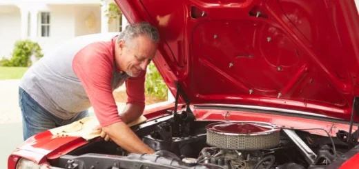 We view an older man learning into the opened hood of a classic car as he works with a wrench. The car hood is read and the man is wearing a light red shirt with a gray vest.
