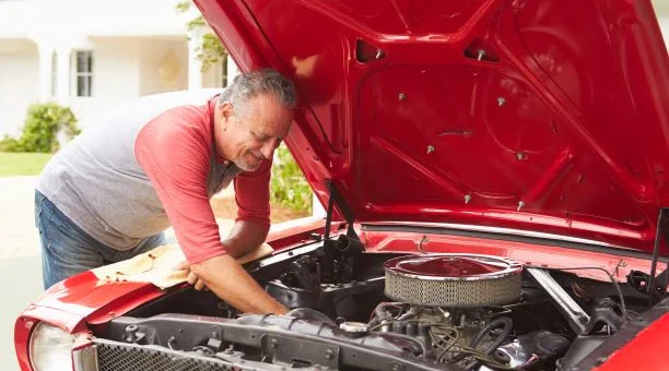 We view an older man learning into the opened hood of a classic car as he works with a wrench. The car hood is read and the man is wearing a light red shirt with a gray vest.