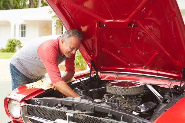 We view an older man learning into the opened hood of a classic car as he works with a wrench. The car hood is read and the man is wearing a light red shirt with a gray vest.
