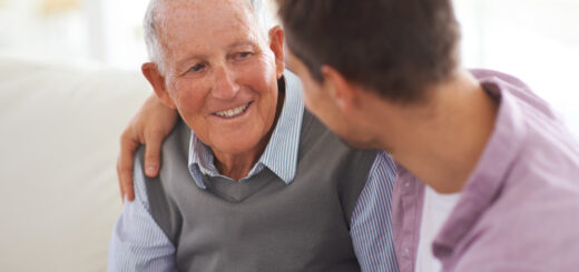 This indoor image shows an older man with short white hair and a receding hairline. He wears a crips light blue collared shirt under a gray pullover sleeveless sweater. Sitting next to him is a younger man who has his arm around the older man's neck as he looks him in the face. We only sewe the back of the younger man's head. He has short brown curly hair and is wearhig a light pinkish colored long sleeved shirt.