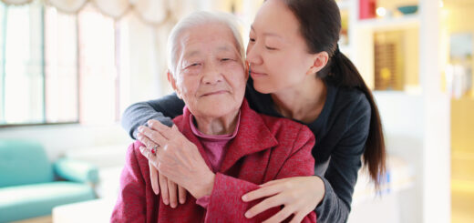 This indoor image shows a woman with Asian features, long, dark hair tied back lovingly hugging an older adult from behind. The older adult, also appearing Asian, has thin gray hair drawn ack and wears a red jacket. The younger woman has one hand on the olde adult's arm and with the oppsing arm slung around their shoulder. The older person is touching the younger person's hand on the shoulder.