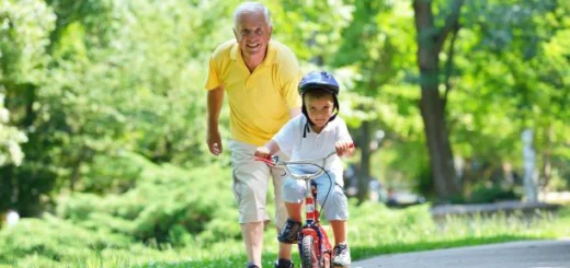 A green park-like view is in the background. On a path in the foreground, we see an older man with gray hair wearing a yellow short-sleeved shirt and beige shorter pants. His hand is on the seat of a small bike as he runs along side while a young child wearing a bike helmet appears to be taking his first ride.
