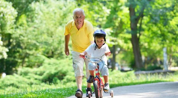 A green park-like view is in the background. On a path in the foreground, we see an older man with gray hair wearing a yellow short-sleeved shirt and beige shorter pants. His hand is on the seat of a small bike as he runs along side while a young child wearing a bike helmet appears to be taking his first ride.