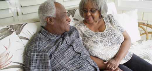 This images shows a couple sitting next to each other on a couch. They look toward each other with loving glances. The man on the left is seen in profile. He has tightly curled, short gray hair receding significantly. He wears glasses and has a gray flannel shirt on. The woman he is look at has short gray hair and wis wearing a gray and white patterned top. Both have dark skin.