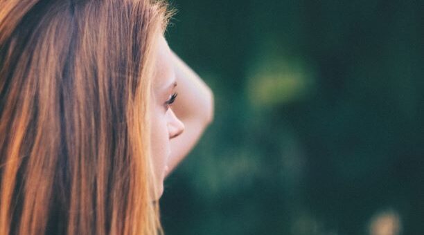 We see a blurred outdoor background with a younger woman in profile. She is looking slightly backward as she raises her hand into her long, red hair as if in frustration.