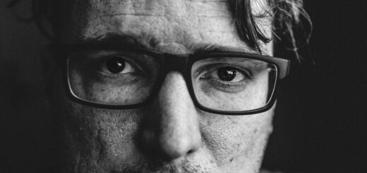 This black and white image shows a closeup of a middle-aged man with short dark hair, a wavy lock falling over his forehead. He wears rectangular, dark rimmed glasses and a worried expression.