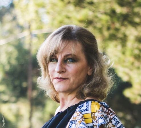 This outdoor image shows a younger woman with long, dark blonde hair. She's turned in profile but slightly faces the camera as she looks to the distance. She wears a patterned blue and yellow blouse over a dark blue apron.