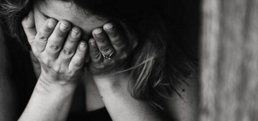 This black and white image shows a woman crying into her hands. Her head is bowed in grief. She has dark hair and medium-dark sking and is wearing a dark top.