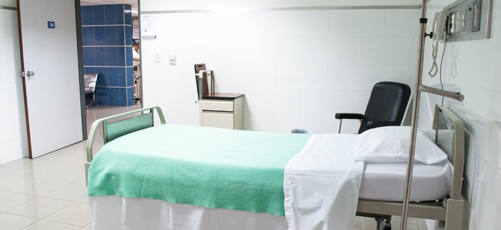 This image shows the interior of a classic hospital room with bed covered in a sheet with a light green blanket. We see a black visitor's chair plus a board for instructions off to the side.